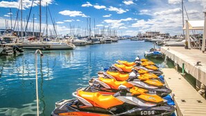 Un puerto deportivo con varias embarcaciones amarradas, una zona de alquiler de motos acuáticas y cielo despejado.