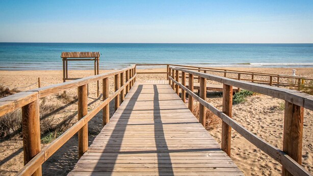 Un muelle de madera que lleva a una playa, con un banco y una torreta de salvavidas.