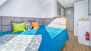 Een moderne slaapkamer met een bed, kussens, een blauwe deken, een plaid met een geometrisch patroon, een wit hoofdbord en een kleine kitchenette met een brandblusser.