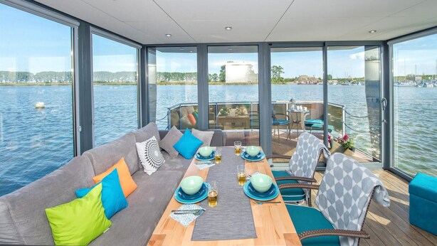 Een ruim terras met een bank, eettafel en stoelen, met uitzicht op een rustig waterlichaam.