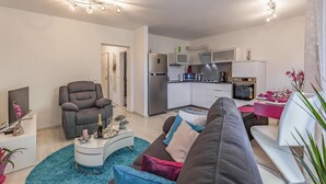 A modern living room with a grey sofa, a white coffee table, and a TV on a stand. A kitchen with white cabinets and stainless steel appliances is visible in the background. - 2 bedroom beautiful apartment in Liznjan (Liznjan)