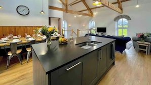 A modern kitchen with a large island, wooden beams, and a dining area with a clock and flowers. - Amazing Chapel near Yr Wyddfa (Snowdon) and Zip World (Llanrug)