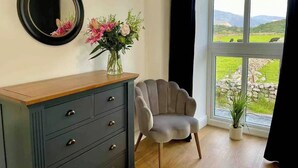 A room with a dresser, a chair, a vase with flowers, a mirror, and a large window with a view of greenery and mountains. - Amazing Chapel near Yr Wyddfa (Snowdon) and Zip World (Llanrug)