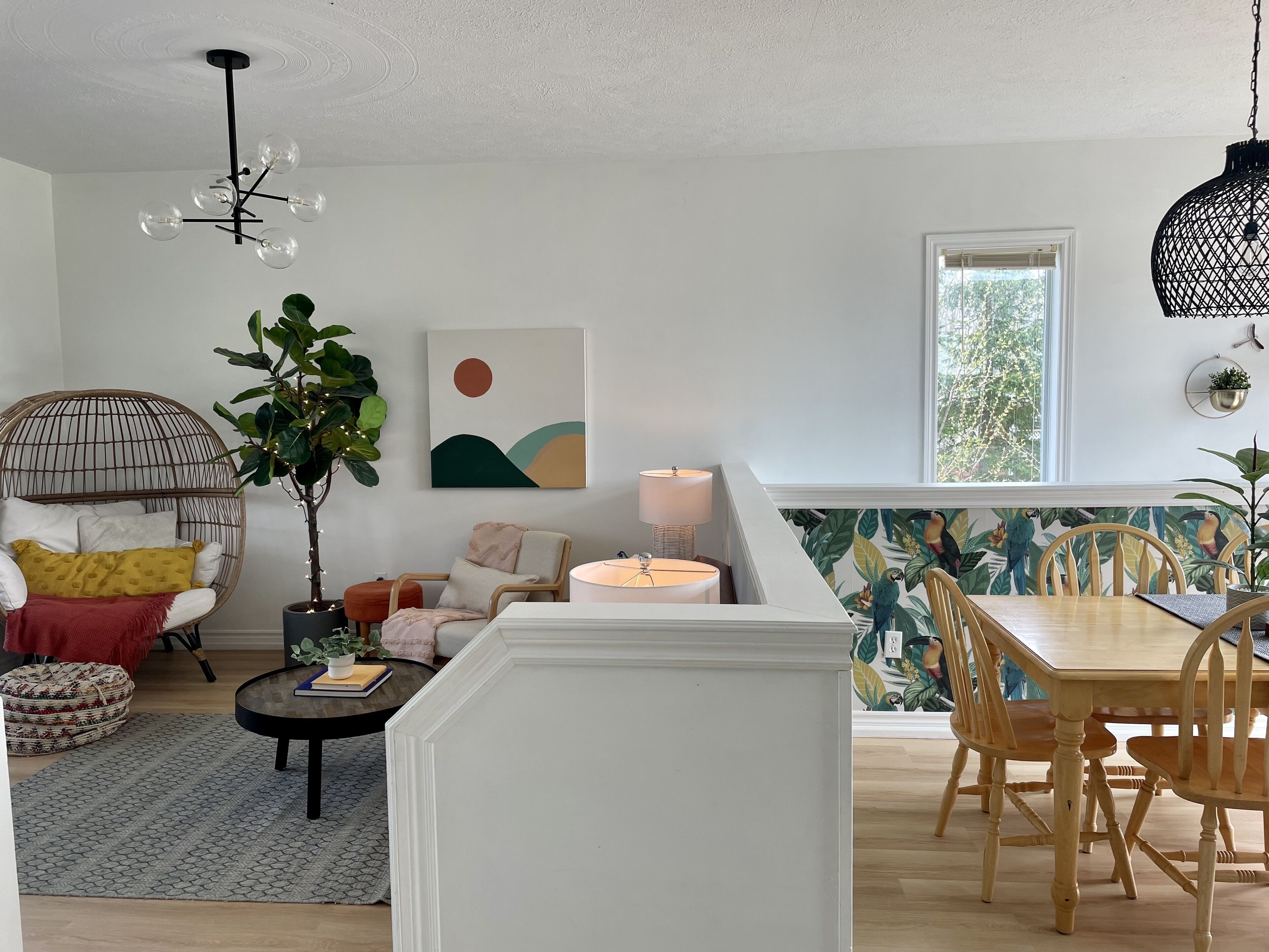 Un salon moderne avec une table basse ronde, un fauteuil en osier, une table à manger avec des chaises en bois, un luminaire suspendu et une grande fenêtre donnant sur les arbres.