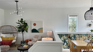 Ein modernes Wohnzimmer mit rundem Couchtisch, einem Korbstuhl, einem Esstisch mit Holzsesseln, einer Pendelleuchte und einem großen Fenster mit Blick auf Bäume.