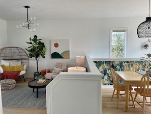 Ein modernes Wohnzimmer mit rundem Couchtisch, einem Korbstuhl, einem Esstisch mit Holzsesseln, einer Pendelleuchte und einem großen Fenster mit Blick auf Bäume.