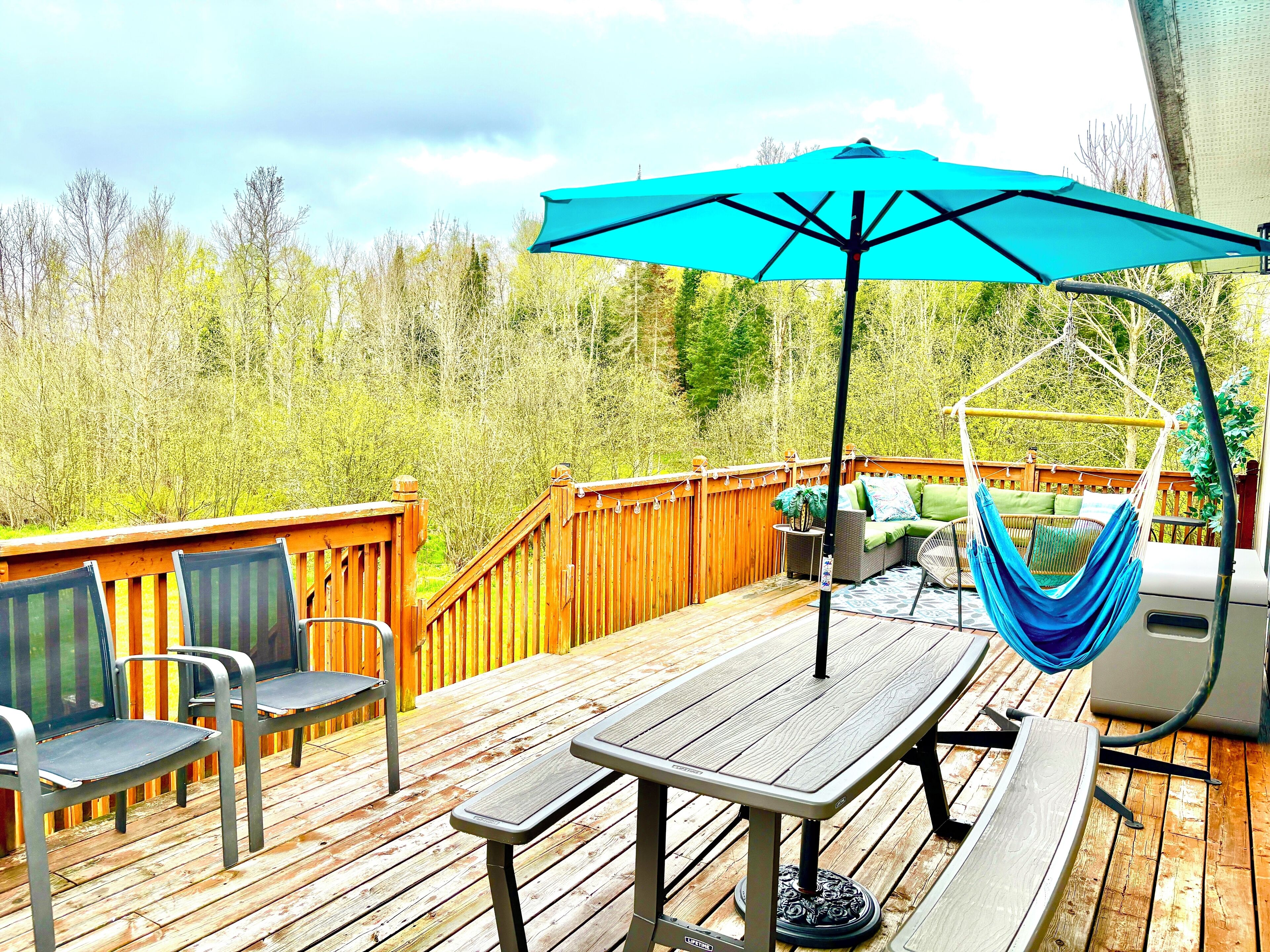 Une terrasse en bois avec une table et des chaises, un hamac et un parasol bleu.