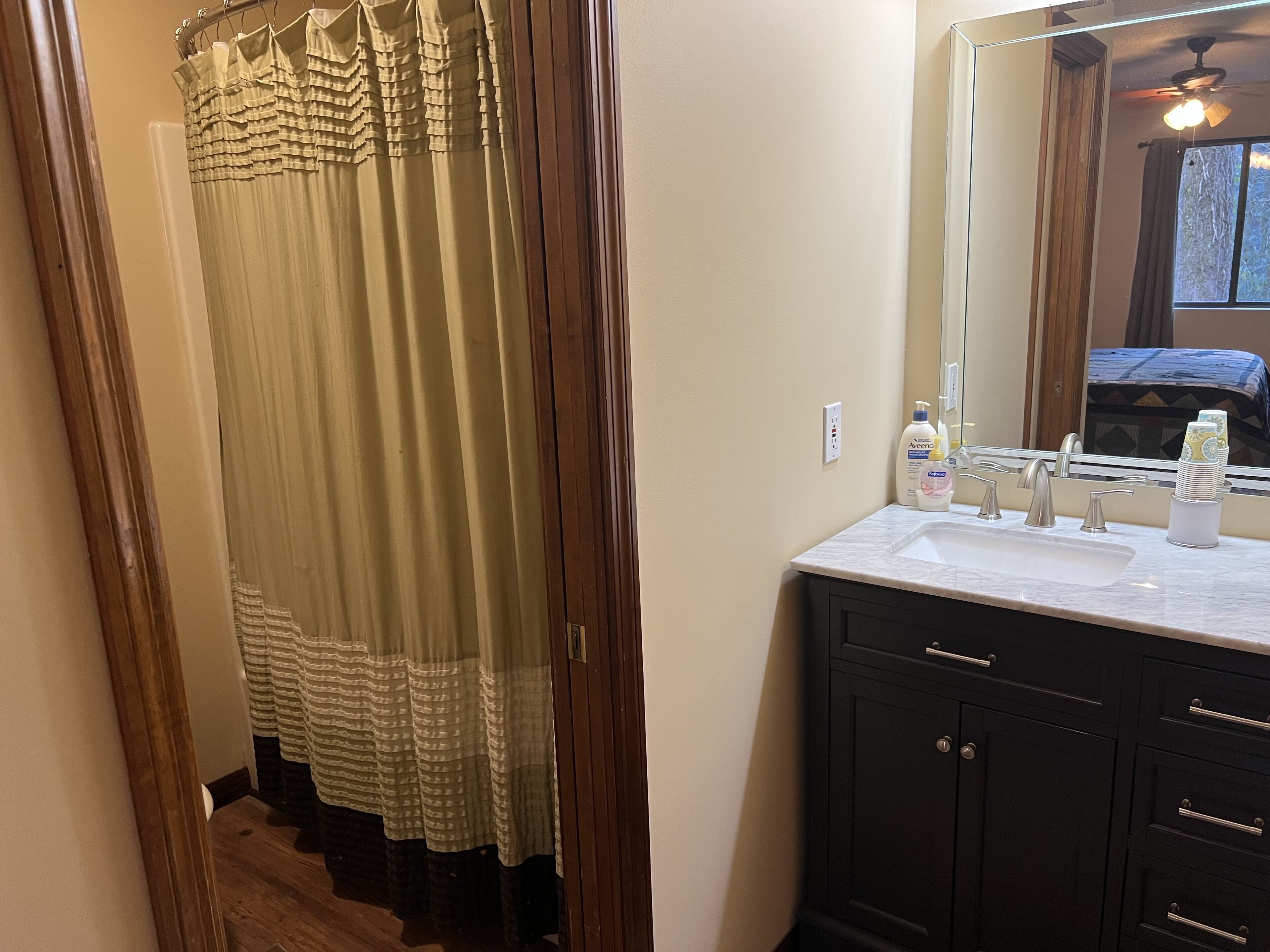 A bathroom with a shower curtain, a sink with toiletries, and a mirror reflecting a bed.