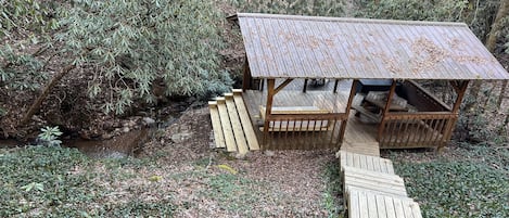 En rustik träkonstruktion med ett tak av metall, omgiven av tät vegetation och med en träveranda som leder fram till den.