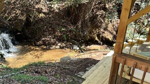 Une terrasse en bois surplombe un ruisseau naturel avec une cascade, le tout étant entouré d’une végétation luxuriante.