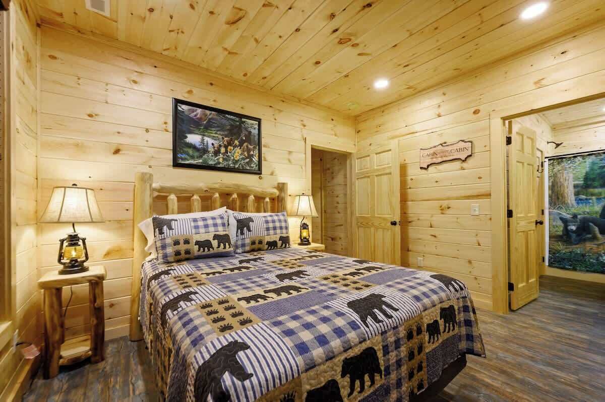A wooden cabin bedroom with a bed, two bedside lamps, a framed picture on the wall, and a door with a bear illustration.