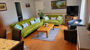 Een woonkamer met een groene bank, een houten salontafel en een zwarte leren fauteuil. Er staat een televisie op een houten dressoir en een boekenkast vol boeken.