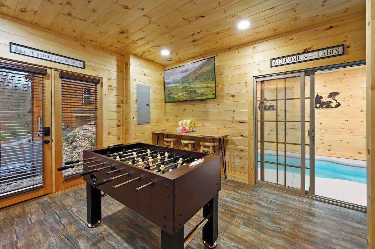 A wooden cabin interior with a foosball table, a television, and a scenic view through the glass door.