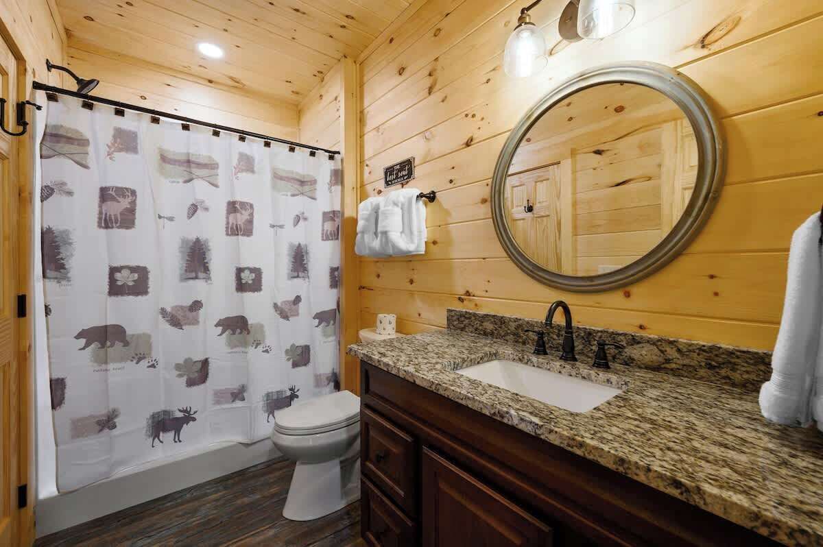 A wooden-paneled bathroom with a shower curtain featuring animal prints, a toilet, a sink with a granite countertop, and a round mirror.
