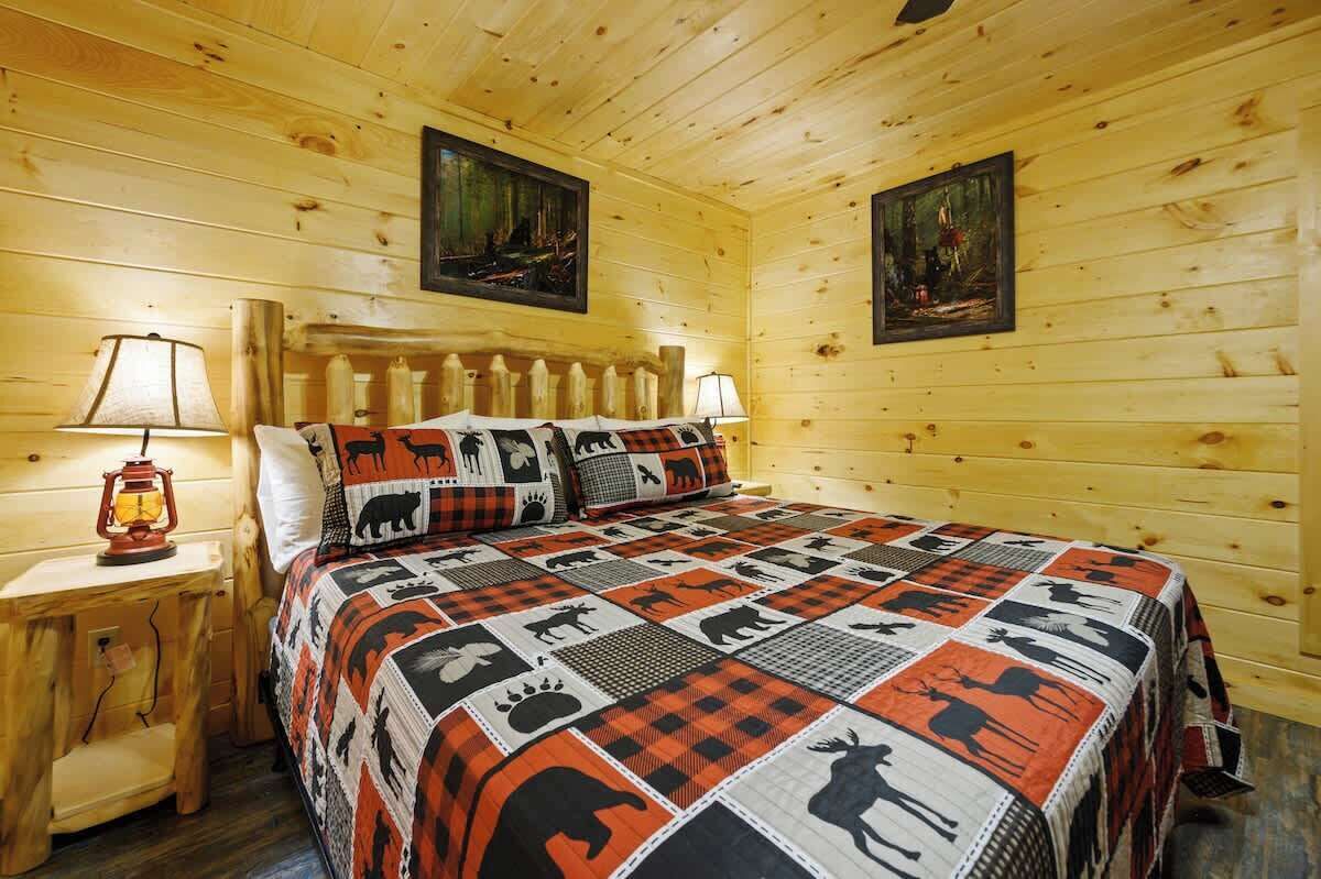 A wooden cabin room with a bed, two paintings, and a nightstand with a lamp.