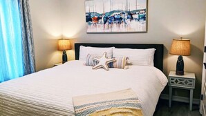 A neatly made bed with a starfish pillow, a painting of boats on the wall, and two bedside lamps. - Howdy Holiday: Modern Family Getaway (Corpus Christi)
