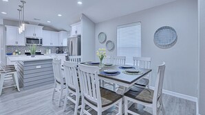 A modern kitchen with white cabinets, a central island, and a dining area with white chairs and a wooden table. - Howdy Holiday: Modern Family Getaway (Corpus Christi)