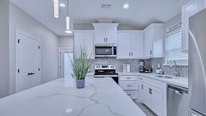 A modern kitchen with white cabinetry, a large marble island, stainless steel appliances, and pendant lighting. - Howdy Holiday: Modern Family Getaway (Corpus Christi)