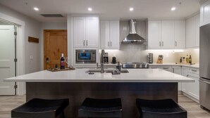 A modern kitchen with white cabinets, a large island with stools, a stainless steel range hood, and a built-in microwave. - Le Bucheron Stunning Spring Creek Luxury condo (Canmore)