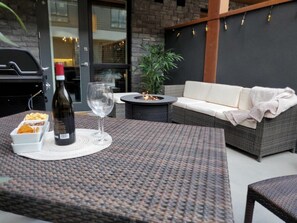 Un área de estar al aire libre con una mesa, una botella de vino y vasos. Hay un brasero, un sofá y una planta en maceta.