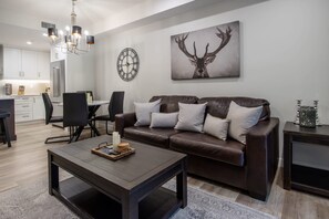 Una sala de estar moderna con un sofá de cuero marrón, una mesa de centro, una zona de comedor con una araña y un reloj de pared.