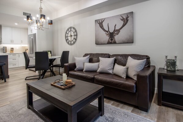 A modern living room with a brown leather sofa, a coffee table, a dining area with a chandelier, and a wall clock. - Le Bucheron Stunning Spring Creek Luxury condo (Canmore)