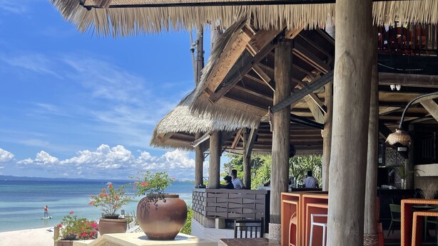 A beachside restaurant with a thatched roof, stone flooring, and wooden furniture.
