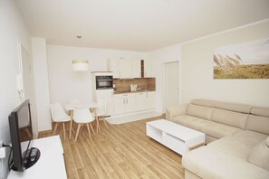 A modern living room with a beige sofa, a white coffee table, and a dining area with a white table and chairs. A kitchen with white cabinets and a wooden countertop is visible in the background.