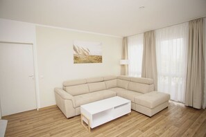 A modern living room with a beige sectional sofa, a white coffee table, and a large window with sheer curtains. - Close to the beach with a terrace; access to the pool and sauna - Strandhaus Mönchgut FeWo06 (Middelhagen)