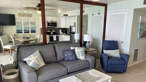 Una sala de estar con un sofá gris, un sillón azul, una mesa de centro blanca y una cocina al fondo.