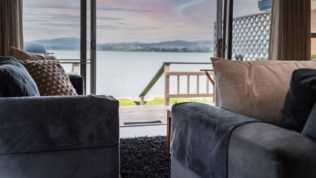 A room with a view of a lake and mountains, furnished with two grey sofas and a wooden table. - Stay Tauranga Beach House (Tauranga)