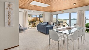 A spacious living area with a glass table, white chairs, a blue sofa, and a view of the sea. - Stay Tauranga Beach House (Tauranga)