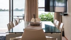 A dining table with chairs, a TV mounted on the wall, and a view of the water outside. - Stay Tauranga Beach House (Tauranga)