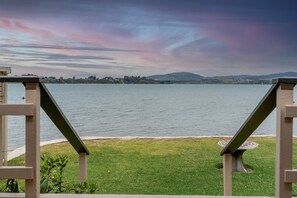 A lakeside view from a balcony with a metal railing, a grassy area, and a distant shoreline with hills.