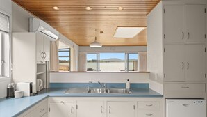 A kitchen with white cabinets, a blue countertop, a sink, and a window with a view of the landscape. - Stay Tauranga Beach House (Tauranga)