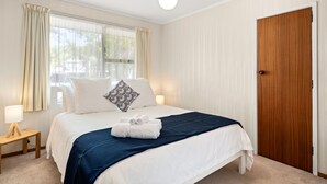 A neatly arranged bedroom with a large bed, a bedside table with a lamp, and a wooden door. - Stay Tauranga Beach House (Tauranga)