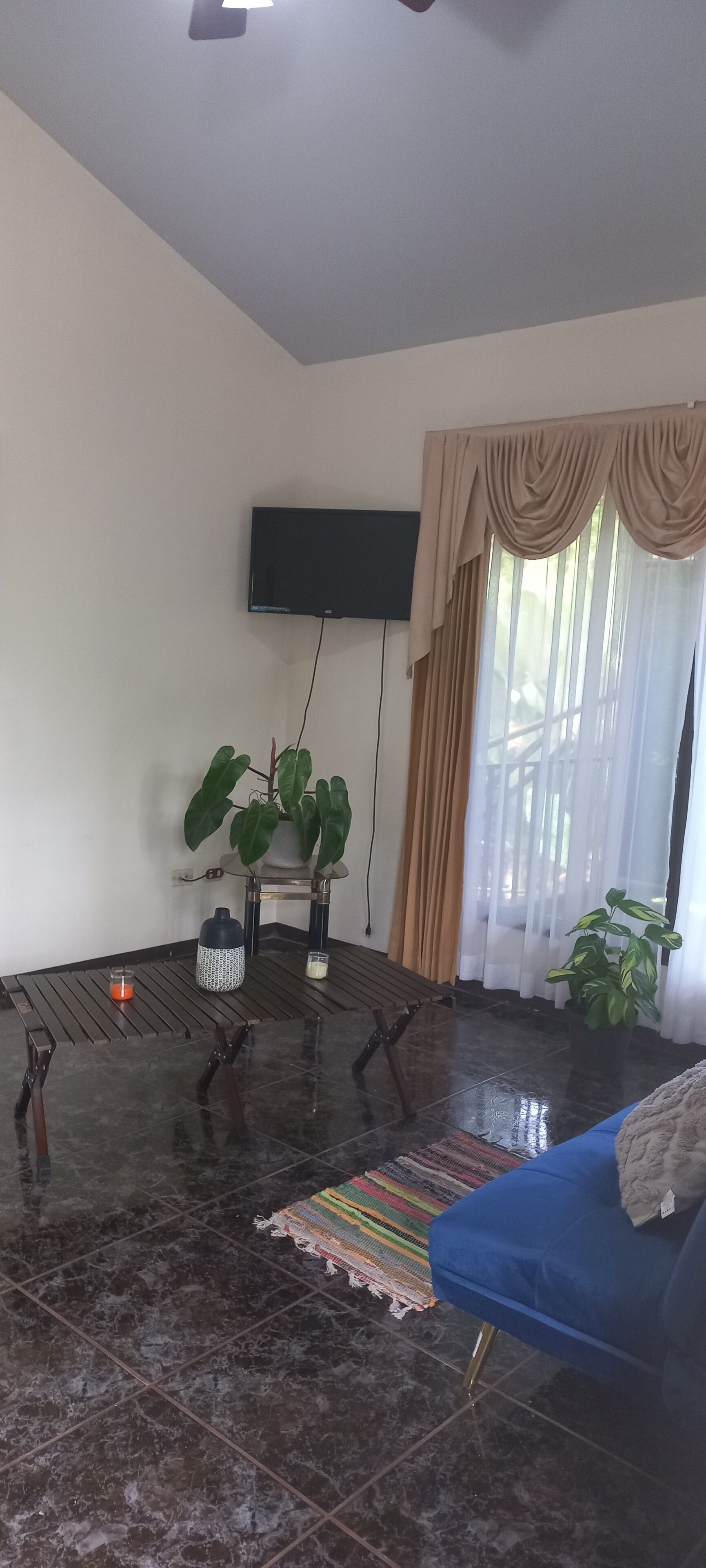 A living room with a dark tiled floor, a blue sofa, a flat-screen TV mounted on the wall, and a large window with sheer curtains.