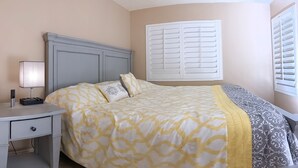Un dormitorio con cama, mesita de noche, lámpara y ventana con contraventanas.