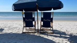 Two beach chairs labeled "Beach Colony" under a large umbrella on a sandy beach with the ocean in the background. - 7th floor 2 bedroom 2 bath oceanfront condo at Beach Colony West. (Navarre)