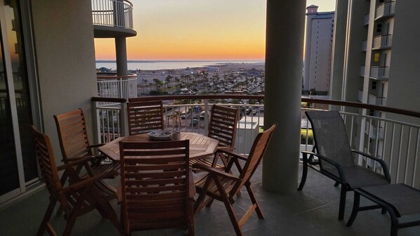 Un balcón con una mesa y sillas, con vistas a un paisaje urbano y al océano al atardecer.