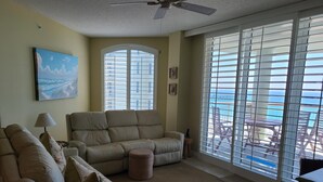 A living room with a beige sofa, a small ottoman, a ceiling fan, and large windows with blinds. - 7th floor 2 bedroom 2 bath oceanfront condo at Beach Colony West. (Navarre)