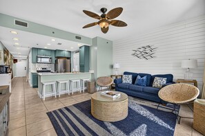 A modern kitchen with a central island, a dining area with white stools, a blue sofa with patterned cushions, and a ceiling fan. - Beachfront Condo at Sterling Breeze - heated pool, hot tub, gym, Wi-Fi (Panama City Beach)