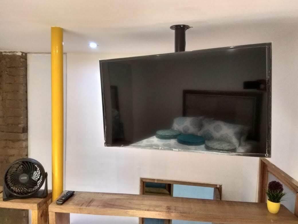 Une pièce dotée d’un grand téléviseur à écran plat fixé au mur, d’une table en bois avec une télécommande, d’un petit ventilateur et d’une plante en pot.