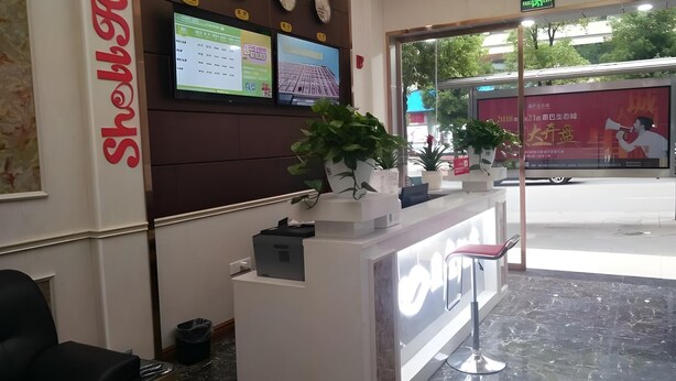 Un comptoir d’accueil d’hôtel doté d’un écran numérique affichant des informations sur les chambres, une horloge et des plantes en pot.