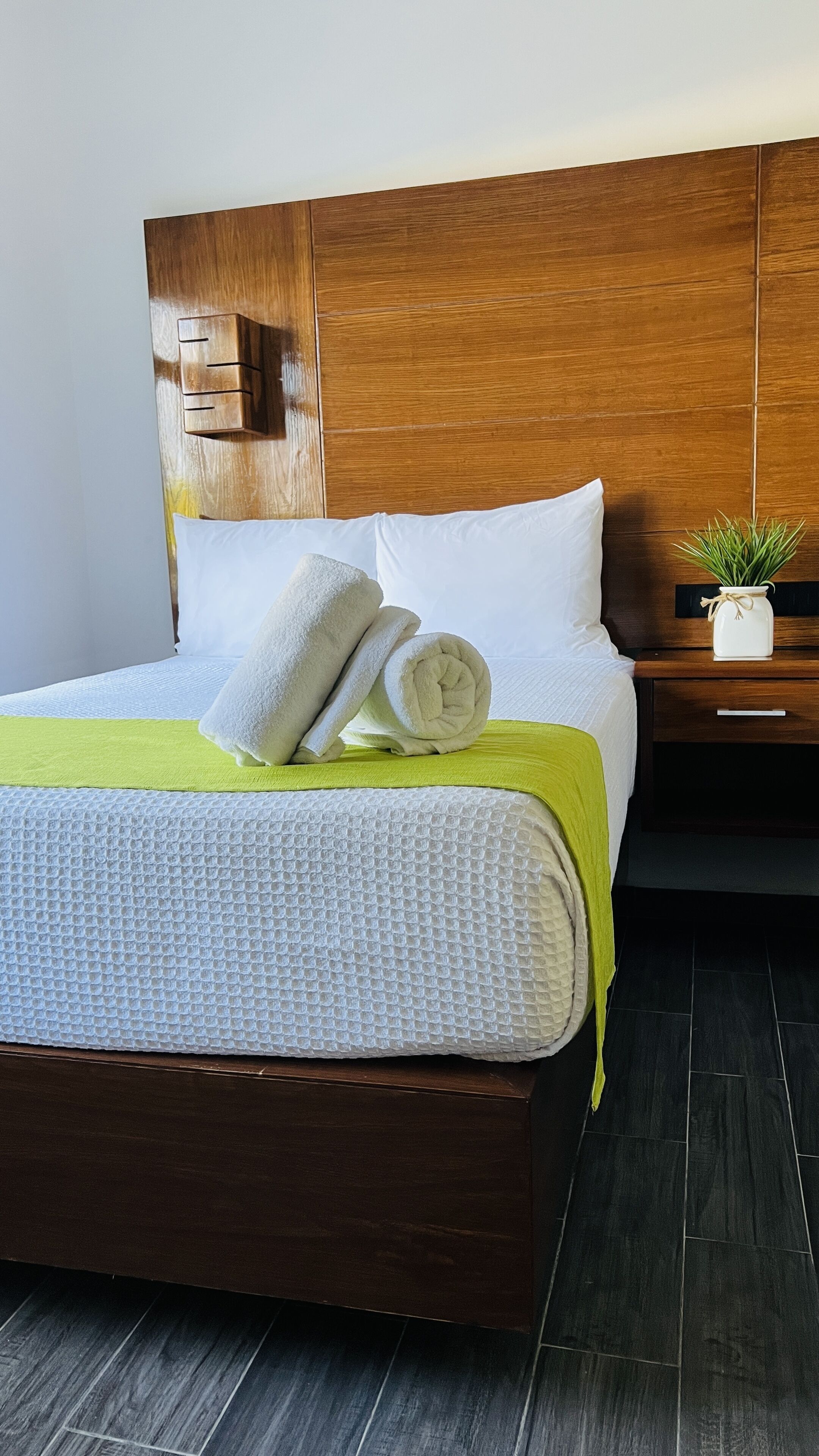 Een net opgemaakt bed met een witte, geribbelde sprei, twee opgerolde witte handdoeken en een groene plaid.