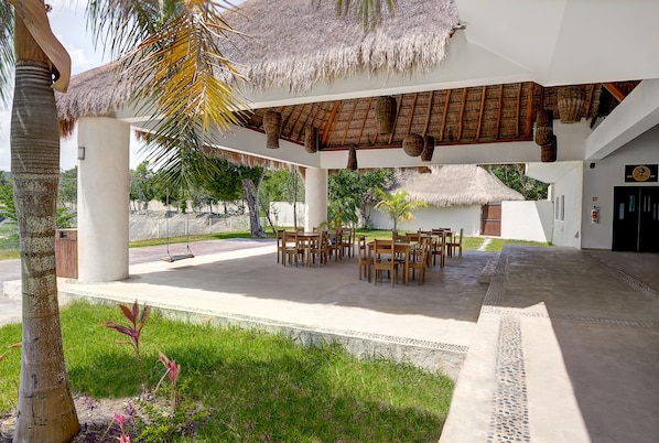 An outdoor seating area with thatched roof and wooden furniture, surrounded by greenery and a palm tree.