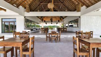 A spacious dining area with wooden tables and chairs, a thatched roof, and large windows offering views of the outdoors.