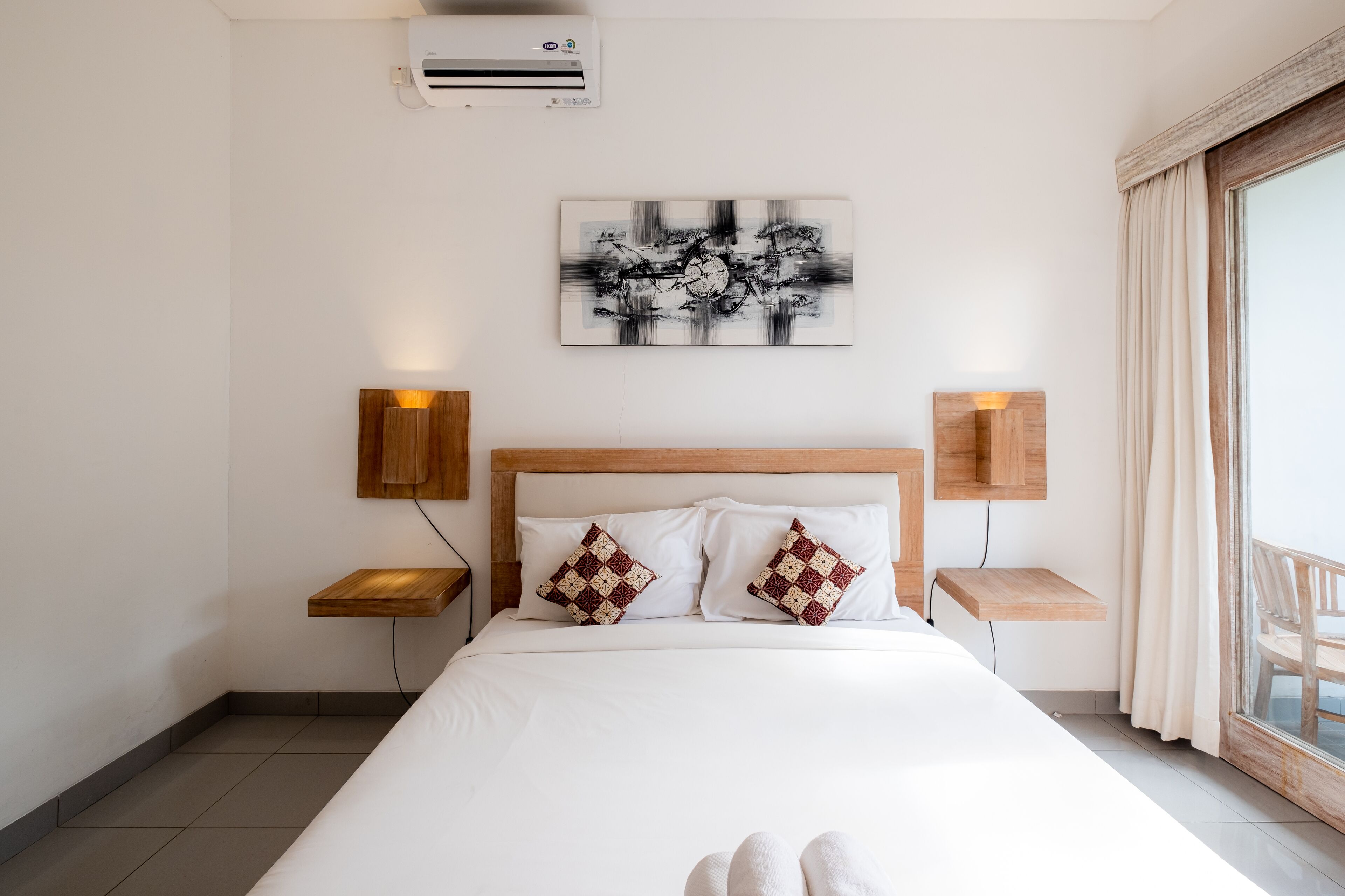 A hotel room with a large bed, two bedside tables with lamps, a wall-mounted air conditioner, and a large window with curtains.