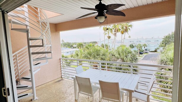 Une terrasse extérieure couverte, dotée d’une table et de chaises, d’un ventilateur de plafond et offrant une vue sur un quartier résidentiel avec des palmiers et des maisons.