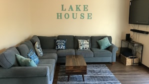 A living room with a grey sectional sofa, a wooden coffee table, and a blue rug. The wall has a sign that reads "LAKE HOUSE". - Lake House w/ Pool and King Bed, Fishing - Dogs OK (Hedgesville)